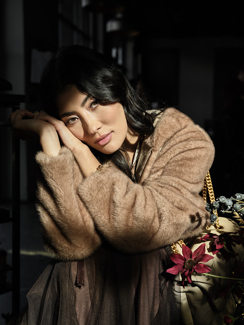 Eine Frau posiert in einer braunen Fake-Fur-Jacke vor der Kamera. Am rechten Bildrand streift ein herbstliches Blumen-Bouquet ihren Look.