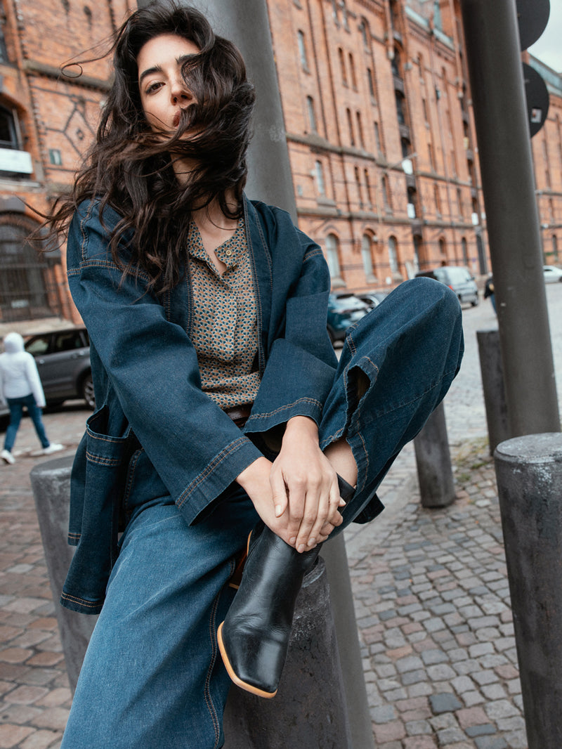 Une femme est assise sur un poteau métallique. Elle porte une longue chemise en jean et un pantalon assorti. Ses cheveux flottent dans le vent hambourgeois.