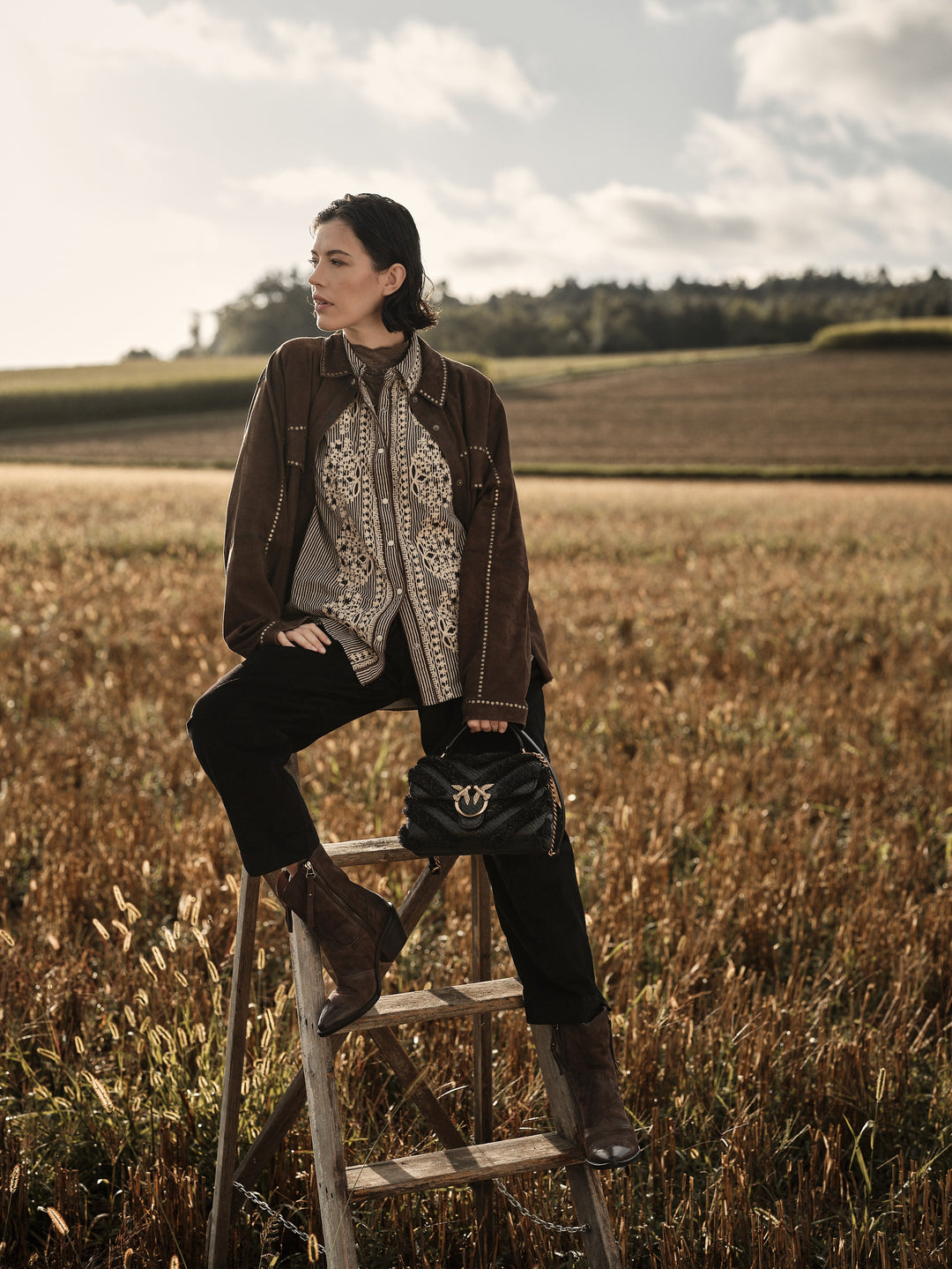 Eine Frau sitzt in einem Feld auf einer Holzleiter. Sie trägt einen Look von Bijou les Boutiques.